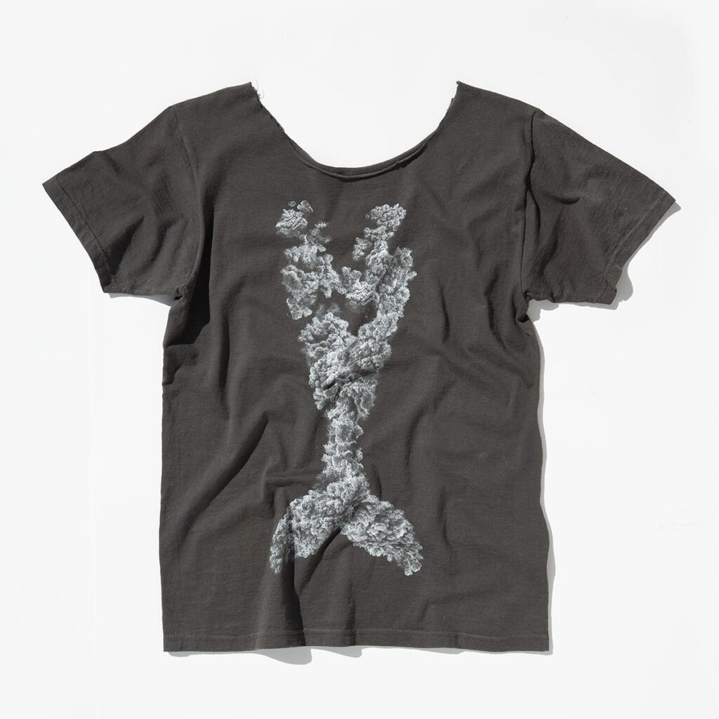 T-shirt scura con un disegno chiaro che rappresenta una coda di balena stilizzata, composta da un motivo simile a un bosco o una formazione naturale.