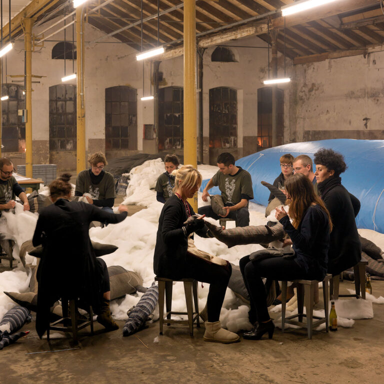 Partecipanti seduti in cerchio attorno alla scultura di una balena, intenti a cucire e lavorare con materiali tessili all'interno di un grande spazio industriale.